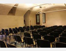 Sala conferenze interna con soffitto a volta e pareti color crema, illuminata da faretti su binari. File di sedie nere disposte in platea di fronte a una parete, con finestre e una porta sul lato.