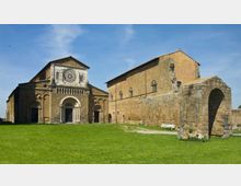Veduta esterna di una chiesa in pietra in stile romanico con grande rosone e portale ad arco, affiancata da lunghi edifici monastici. In primo piano un prato verde e, sulla destra, un arco in muratura con resti di strutture in pietra sotto cielo sereno.