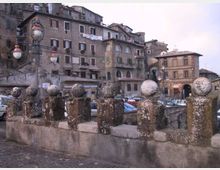 Balaustra in pietra con pilastrini sormontati da sfere in primo piano, lungo una piazza lastricata. Sullo sfondo si vedono edifici storici addossati su un pendio, con facciate in muratura, finestre e tetti in tegole, oltre a lampioni e auto parcheggiate.