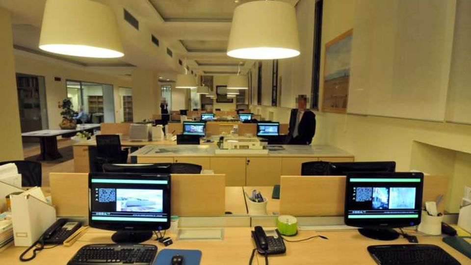 Interno di un ufficio open space con file di scrivanie in legno chiaro dotate di computer e telefoni, illuminate da grandi lampade a sospensione. Sullo sfondo si vedono pareti chiare, un corridoio centrale e alcune vetrate e quadri alle pareti.