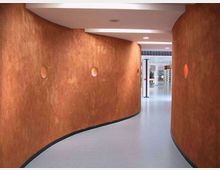 Corridoio interno moderno con pareti curve color terracotta e piccoli oblò circolari incassati, pavimento grigio chiaro e soffitto bianco con luci integrate. Il passaggio sinuoso conduce a un’area più ampia e luminosa visibile in fondo.