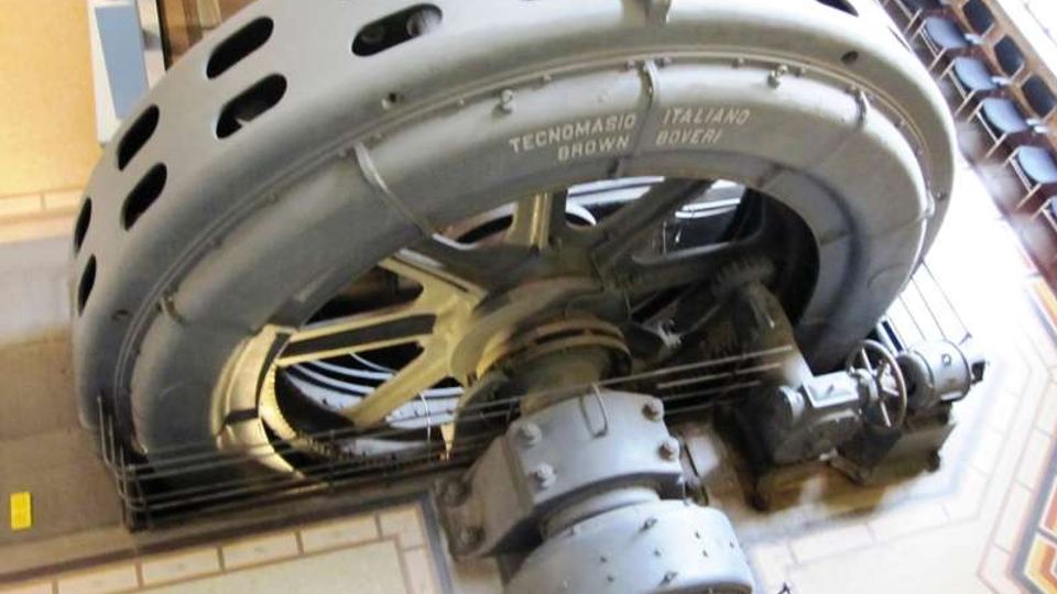 Grande macchinario industriale d’epoca con una massiccia struttura circolare metallica forata e componenti meccanici interni visibili. L’attrezzatura è installata in un ambiente interno con pareti e finiture architettoniche, simile a un museo o a un edificio storico.