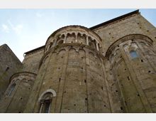 Vista dal basso dell’abside di una chiesa in pietra in stile romanico, con volumi semicircolari e fasce di archetti ciechi. In alto si notano una loggia ad arcate con colonne e piccole finestre inserite nei muri spessi.