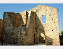 Ruderi di un edificio in pietra con muri alti e aperture senza infissi, tra cui una grande finestra rettangolare e un arco di passaggio al piano terra. La struttura è priva di tetto e si trova all’aperto sotto un cielo sereno.