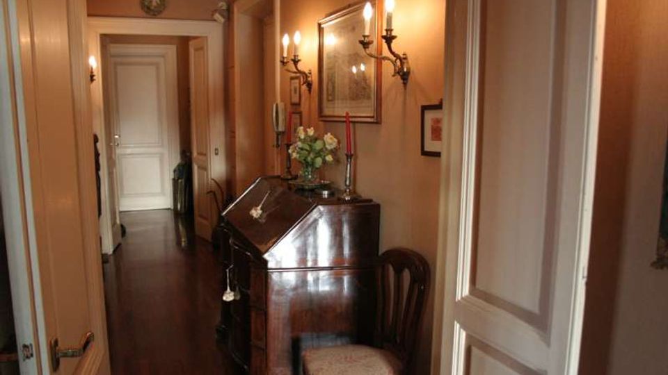 Corridoio interno stretto con pareti color beige, pavimento in legno scuro e porte bianche lungo il passaggio. Sul lato destro ci sono applique a parete, quadri e un mobile scrittoio in legno con fiori e candelabri, affiancato da una sedia.