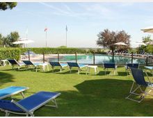 Piscina all’aperto circondata da prato con numerosi lettini blu e ombrelloni bianchi. Intorno si vedono siepi, alcuni alberi e pennoni con bandiere sullo sfondo sotto un cielo sereno.