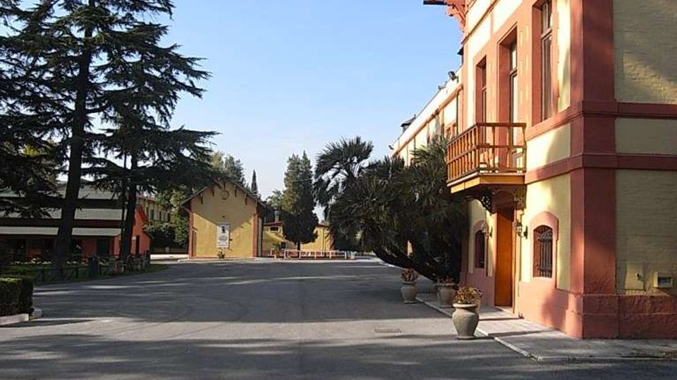 Cortile o viale asfaltato in un complesso con edifici bassi; a destra una palazzina color crema e rosso con balconcino in legno e tetto sporgente. L’area è circondata da alberi e piante in vaso sotto un cielo sereno.