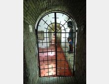 Corridoio sotterraneo con volta in mattoni e pavimento rossastro, visto attraverso un cancello in ferro ad arco. In fondo si intravedono pareti bianche illuminate e una serie di passaggi ad arco lungo il tunnel.