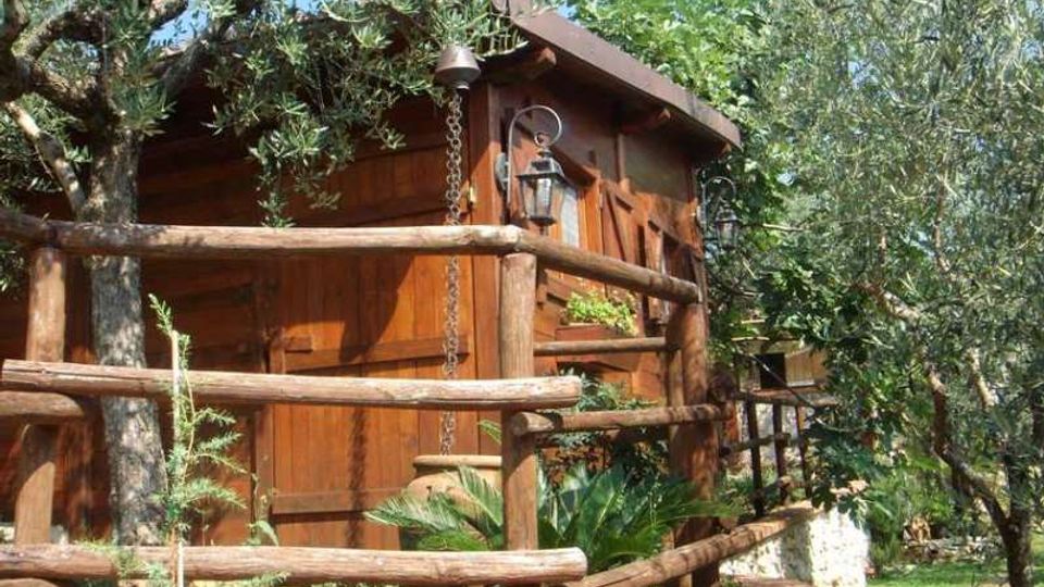 Casetta in legno con tetto spiovente, circondata da alberi e vegetazione, con staccionata in tronchi e piccola aiuola rialzata in mattoni. Sulla facciata sono visibili una lanterna a parete e una catena pluviale.