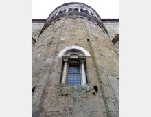 Vista dal basso di una torre cilindrica in pietra di un edificio storico, con piccola finestra ad arco incorniciata da due colonne e grata. In alto si nota un cornicione con archetti decorativi lungo il bordo.