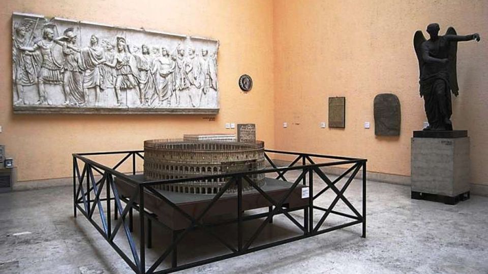 Sala museale con pareti color ocra e pavimento in pietra, con al centro un grande modello circolare recintato da una struttura metallica. Sulle pareti sono esposti un lungo rilievo marmoreo e alcune lapidi, con una grande statua scura su piedistallo in un angolo.