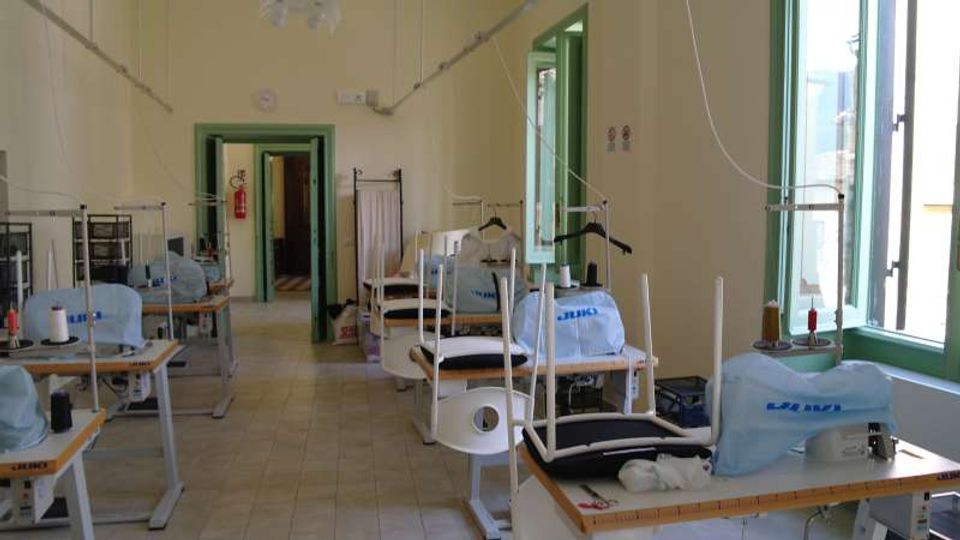 Ampio laboratorio di sartoria con file di tavoli da lavoro dotati di macchine da cucire e supporti per filo, pavimento piastrellato e pareti chiare. Grandi finestre con infissi verdi lungo il lato destro illuminano l’ambiente, con una porta sul fondo che conduce a un corridoio.