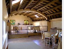Interno di una sala tipo ristorante con soffitto mansardato in legno e travi a vista, pareti chiare e pavimento in legno. Tavoli apparecchiati e sedie sono disposti su due livelli, separati da un gradino e da una ringhiera metallica, con piccoli lucernari sul tetto.