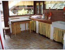 Cucina rustica con grandi finestre in legno che affacciano sul verde, pavimento in piastrelle e piano di lavoro in legno lungo la parete. Sotto il piano e il lavello ci sono tendine gialle, con paraschizzi in mattonelle e utensili da cucina appoggiati sul bancone.