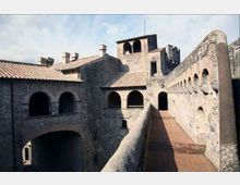 Cortile interno di un edificio storico in pietra con tetti in tegole, arcate su più livelli e un camminamento sopraelevato con parapetto e pavimento in cotto. Sulla destra si vede un lungo muro con una fila di piccole aperture e un accesso ad arco sul fondo.