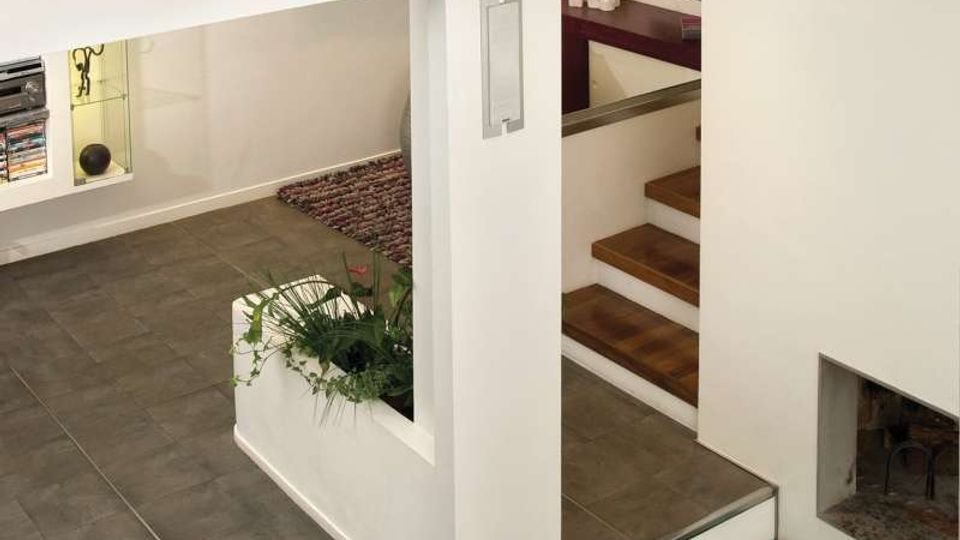 Interno di una casa moderna con pareti bianche e pavimento in grandi piastrelle grigie, organizzato su più livelli con gradini rivestiti in legno. In primo piano si vede un canale incassato con copertura in vetro trasparente e oggetti decorativi, accanto a una scala e a nicchie con piante e mensole.