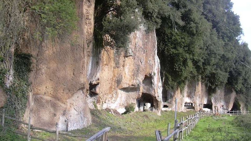 Sentiero erboso con staccionate in legno che conduce a una parete rocciosa tufacea, punteggiata da piccole cavità e aperture simili a grotte. In alto la scarpata è coperta da alberi e vegetazione, con prato ai piedi della roccia.