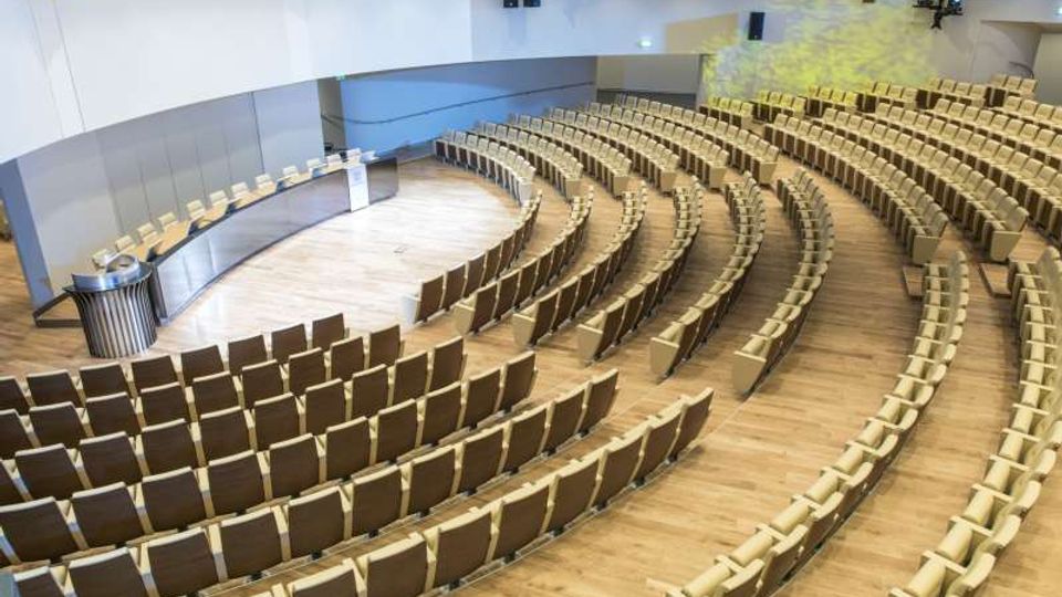 Interno di un’aula magna moderna con pavimento in legno, file di sedute disposte a semicerchio e una postazione relatori con tavolo e podio. Pareti chiare con pannelli acustici, illuminazione da soffitto e balconata laterale sopraelevata.