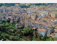 Veduta aerea di un borgo storico su pendio con case in pietra addossate e tetti in coppi, attraversato da strade strette. Al centro spicca una chiesa con cupola e campanile, circondata da vegetazione e terreni verdi ai margini dell’abitato.