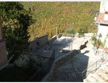 Terrazza in pietra su un pendio, delimitata da muretti e ringhiere, con pavimentazione in lastre e un piccolo spazio recintato. Sullo sfondo si vede una valle boscosa e una collina, con parte di un edificio dai tetti in coppi sulla destra.