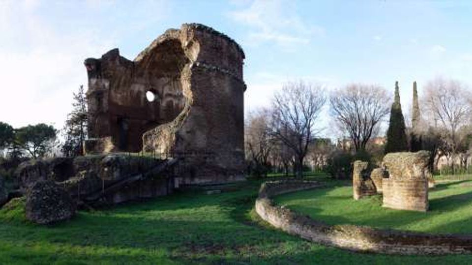 Panorama di un’area archeologica con grandi rovine in mattoni di un edificio antico e diversi resti di muri e pilastri sparsi su un prato. Sullo sfondo si vedono alberi e cipressi sotto un cielo sereno.