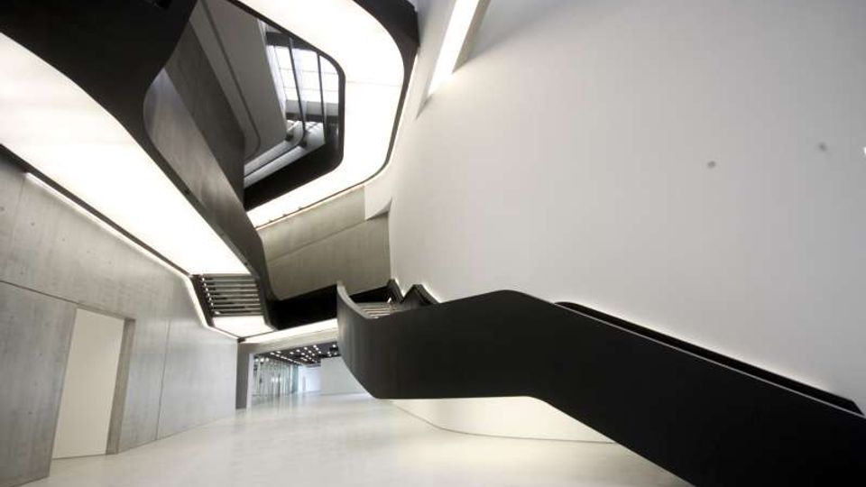 Atrio interno moderno e minimalista con ampie pareti bianche e in cemento e pavimento chiaro. Una scala nera scultorea con linee curve sale verso un lucernario superiore, con passerelle e aperture su più livelli.