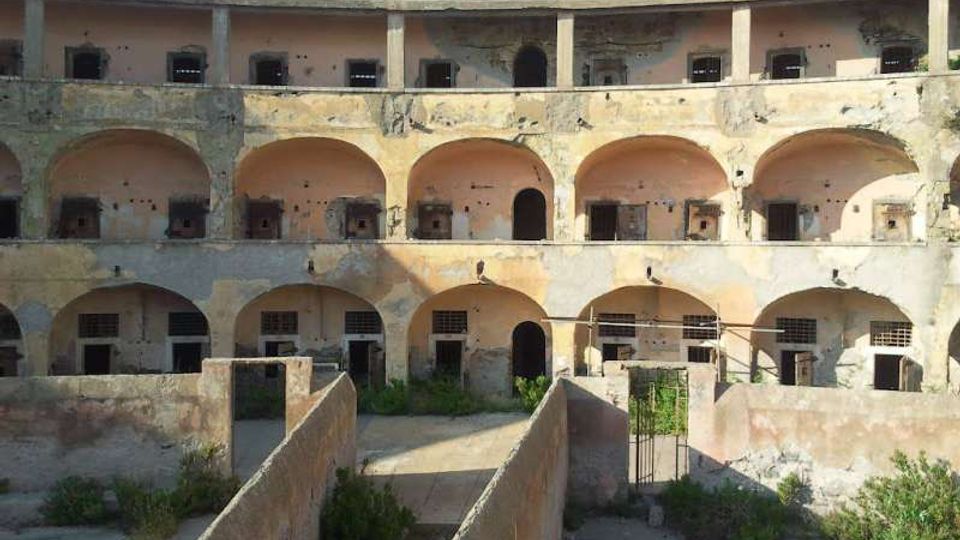 Cortile interno di un edificio storico in rovina, con struttura semicircolare a più piani di arcate e aperture murate o sbarrate. In primo piano si vedono muretti bassi che delimitano corridoi e aree con vegetazione spontanea.