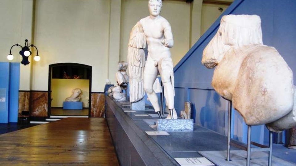 Sala museale con pavimento in legno e pareti chiare, con una lunga pedana espositiva che ospita statue e frammenti scultorei in marmo. In primo piano si vedono un torso e una statua classica, mentre sullo sfondo sono presenti un lampione a più luci e un’apertura verso un’altra sala.