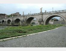 Ponte storico in pietra con più arcate che attraversa un fiume, con statue lungo la balaustra e lampioni. In primo piano si vede una strada acciottolata con una fascia erbosa lungo la riva, e sullo sfondo edifici urbani.