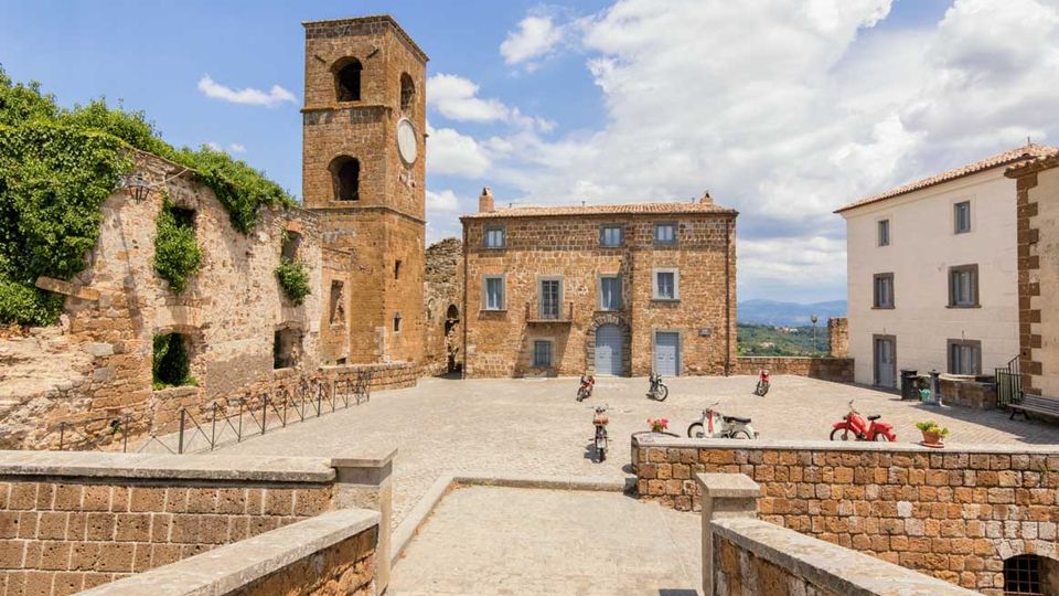 Piazza lastricata in un borgo storico con edifici in pietra e una torre campanaria quadrata con orologio; a sinistra si vedono muri diroccati coperti di vegetazione. Sullo sfondo compaiono case con tetti in coppi e un panorama collinare sotto un cielo parzialmente nuvoloso.