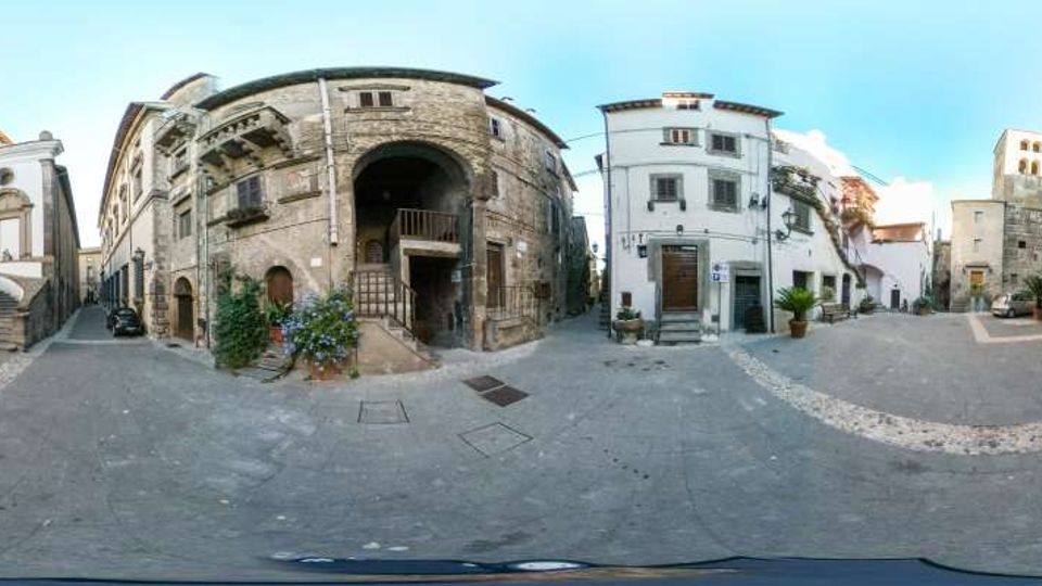 Piazza lastricata in un centro storico, circondata da edifici in pietra e intonaco con archi, scale esterne e balconi. Sullo sfondo si vede una torre campanaria e stradine laterali che si aprono tra le facciate.