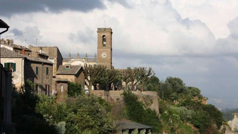 Veduta di un borgo collinare con edifici in pietra e un campanile con orologio che svetta al centro. In primo piano si vede vegetazione fitta e una terrazza murata con alberi potati, sullo sfondo cielo nuvoloso e paesaggio in lontananza.