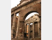 Rovine di una struttura romana con grande arco in mattoni e colonne in pietra scanalate, alcune con capitelli decorati. Sullo sfondo si vedono muri parzialmente crollati e un edificio moderno, con recinzioni metalliche in primo piano.