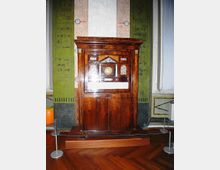 Mobile antico in legno lucido con vetrina, simile a una pendola o a un espositore con quadrante e decorazioni interne, collocato su una pedana e circondato da cordoni di protezione. Sullo sfondo si vede una parete decorata con pannelli verdi con scritte e motivi ornamentali e un pavimento in parquet.