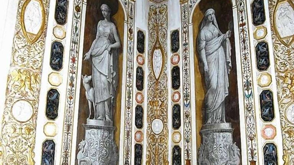 Interno di una sala decorata con stucchi dorati e pannelli ornamentali, con due grandi nicchie ad arco contenenti figure classiche in rilievo. Le pareti sono arricchite da cornici e medaglioni dipinti con motivi naturalistici lungo la parte inferiore.