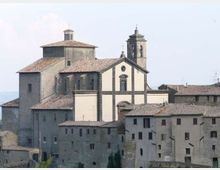 Chiesa in muratura su un borgo collinare, con facciata chiara incorniciata da pietra scura e campanile quadrato con aperture ad arco. Intorno si vedono edifici addossati con tetti in coppi e poche finestre, sullo sfondo cielo chiaro.