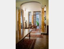 Interno di una sala in stile classico con grandi colonne marmorizzate e arco decorato, pavimento in parquet e tappeti orientali. Al centro un tavolino e, sul fondo, ampie porte a vetri con tende leggere che si aprono su un terrazzo con piante.