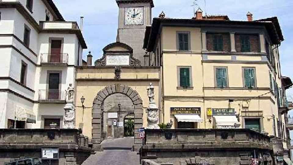 Piazza cittadina con rampa in pavé che conduce a un grande arco in pietra inserito in un edificio storico, con torre dell’orologio sullo sfondo. Intorno si vedono palazzi a più piani con persiane verdi e insegne di negozi, oltre a muretti in pietra e una piccola fontana in primo piano.