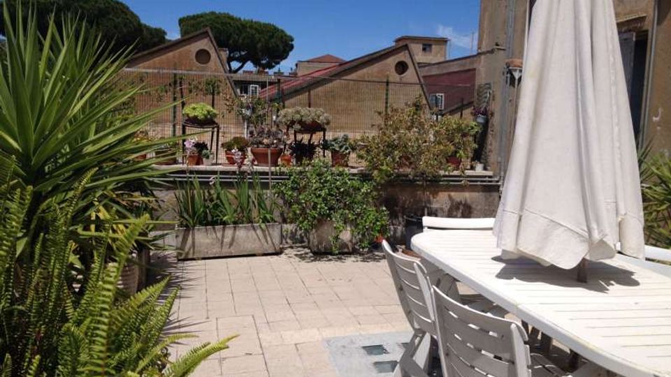 Terrazza pavimentata con tavolo e sedie bianche e un grande ombrellone chiuso, circondata da fioriere e piante in vaso lungo una ringhiera. Sullo sfondo si vedono tetti di edifici e alberi ad alto fusto sotto un cielo sereno.