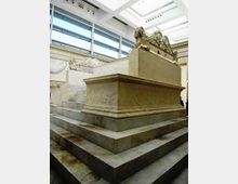 Interno di museo luminoso con grandi finestre e soffitto a lucernario, dove un grande basamento a gradoni in pietra sostiene un sarcofago monumentale scolpito con motivi ornamentali e figure. Pareti in pietra chiara con rilievi decorativi lungo i lati.