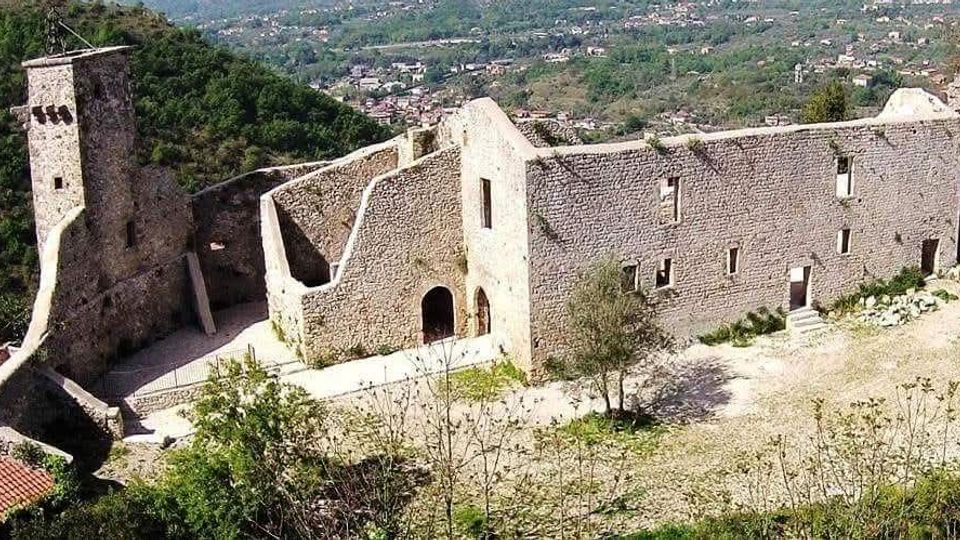 Rovine di un castello in pietra su un pendio, con una torre quadrata, alte mura con piccole finestre e un cortile interno in parte aperto. Sullo sfondo si vede una valle verde con un centro abitato e colline circostanti.