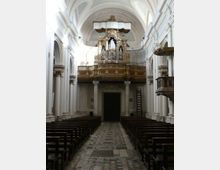 Interno di una chiesa con navata centrale e banchi in legno ai lati, pavimento a mosaico e alte arcate bianche. In fondo si vede una cantoria sopraelevata con un grande organo a canne decorato in oro.