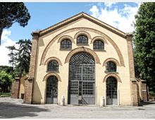 Facciata di un edificio in stile romanico con intonaco chiaro e cornici in mattoni, caratterizzata da grandi archi sovrapposti e ampie finestre ad arco con vetri a griglia. L’ingresso centrale è più alto e vetrato, e l’edificio è circondato da alberi e da un piazzale asfaltato.