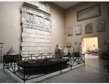Sala museale con pareti color sabbia che espone un grande rilievo lapideo monumentale con fregi scolpiti, affiancato da statue e pannelli in pietra. Al centro c’è una struttura rettangolare recintata in metallo e, sul fondo, un passaggio aperto conduce a un’altra galleria.