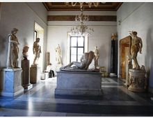 Sala museale con pavimento in marmo a riquadri e soffitto decorato con lampadari, con un grande gruppo scultoreo su piedistallo al centro. Lungo le pareti sono disposte statue classiche su basamenti, con una grande finestra sul fondo e una porta monumentale laterale.