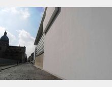 Strada urbana in acciottolato affiancata da un grande edificio moderno con lunga facciata bianca liscia e fascia di finestre orizzontali. Sul lato opposto si vede un edificio storico in ombra con cupola, con cielo parzialmente nuvoloso sullo sfondo.