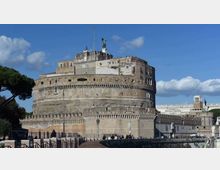Grande fortezza cilindrica in muratura con bastioni e merlature, affacciata sul lungofiume con una strada e recinzioni in primo piano. Sullo sfondo si vedono edifici adiacenti e un cielo azzurro con nuvole.