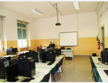 Aula informatica con banchi disposti a isole e postazioni con computer e monitor; sul fondo c’è una lavagna bianca e un proiettore a soffitto. Pareti bicolore bianco e giallo, finestre laterali con tapparelle e illuminazione a neon.
