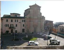 Veduta di una piazza urbana con piccola rotonda centrale erbosa e strada con auto parcheggiate, di fronte a una chiesa storica con facciata barocca in muratura chiara e portale centrale. Intorno si trovano edifici residenziali a più piani con finestre e tetti in tegole, illuminati da luce diurna.