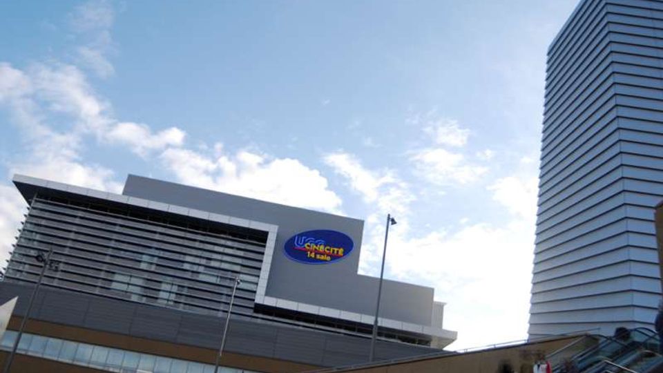Edificio moderno di un complesso commerciale con facciata grigia a pannelli e ampie vetrate, con un grande logo ovale “UCI Cinecittà” in evidenza. A destra si vede una torre alta rivestita da fasce orizzontali e, in primo piano, una scalinata esterna con parapetti e lampioni sotto un cielo parzialmente nuvoloso.